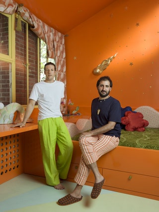 Julien Sebban  seated on bed with green bedding set in an orange frame orange walls Wray standing with hand on orange desk